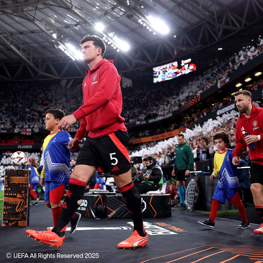 Futbolininkai raudonais marškinėliais ir juodais šortais eina aikštele su vaikais, vilkinčiais sportinę aprangą.