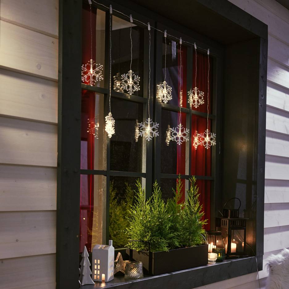 Sniego snaigių girlianda lange su mažais eglutėmis ir žvakėmis ant palangės.