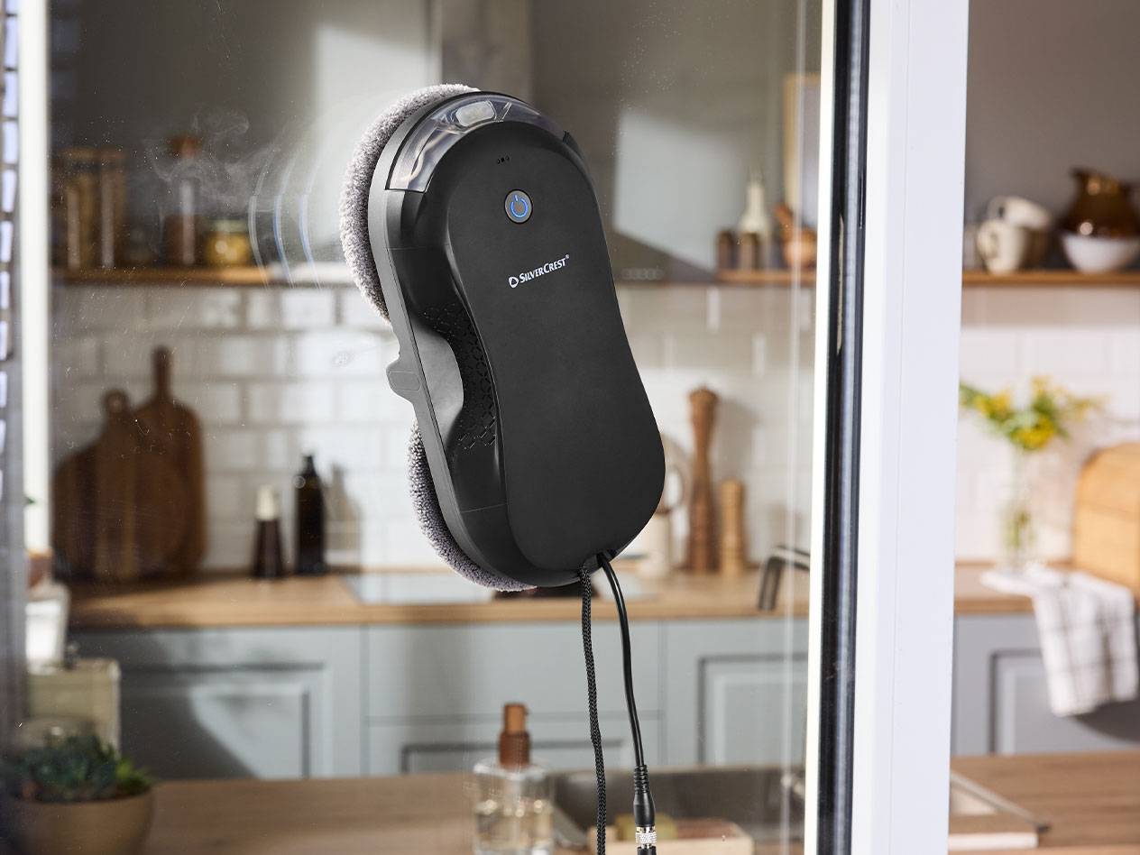 Silvercrest langų valymo robotas ant lango stiklo su virtuve fone.