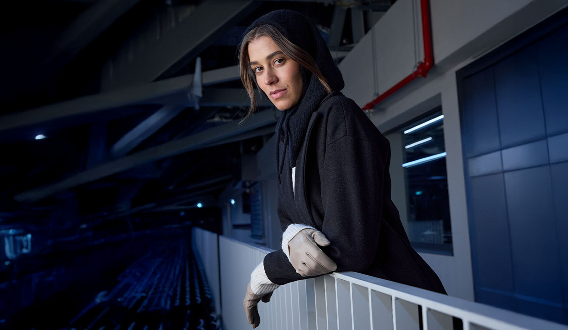 Moteris juodu paltu ir gobtuvu, besiremianti į turėklą modernioje aplinkoje.