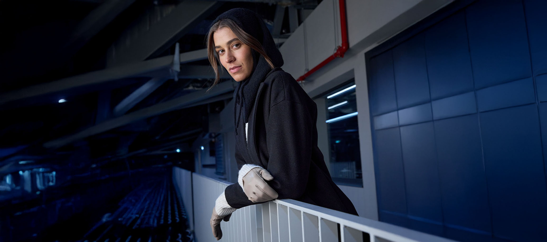 Moteris juodu paltu ir pirštinėmis, pasilenkusi ant turėklų miesto aplinkoje.