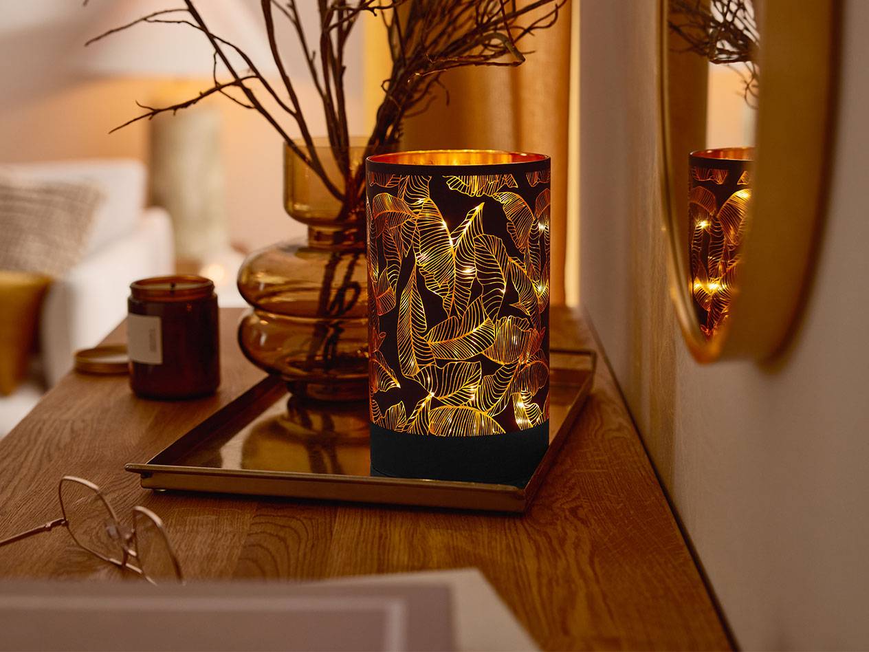 Dekoratyvinis LED žibintas su lapų raštu, vaza su šakomis ir žvakė ant medinio stalo.