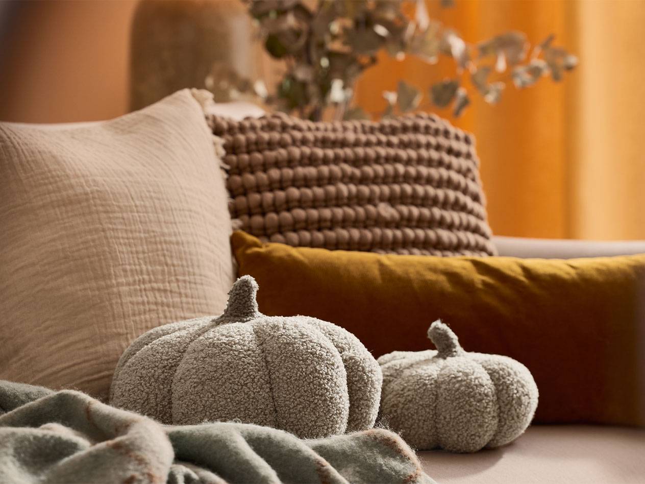 Dvi pliušinės dekoratyvinės moliūgų pagalvėlės ir pagalvės žemės tonų ant sofos.