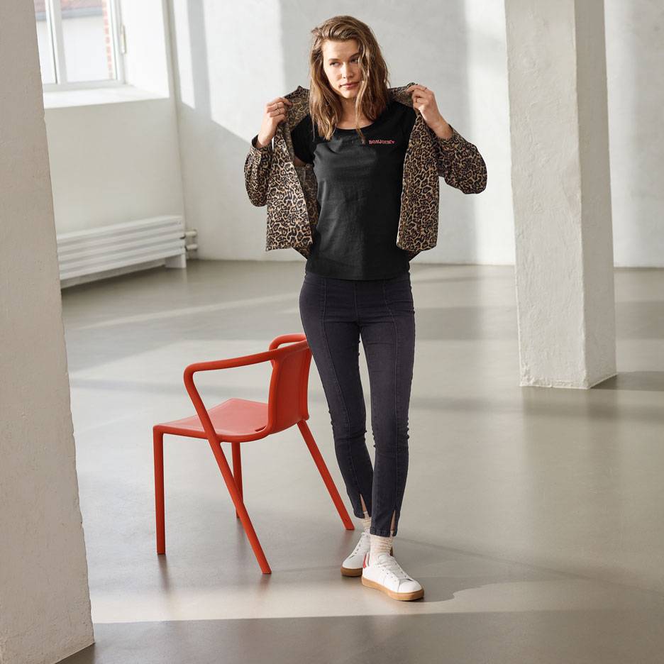 Moteris su juodais marškinėliais su tekstu, džinsais ir leopardo rašto striuke, stovinti prie raudonos kėdės.