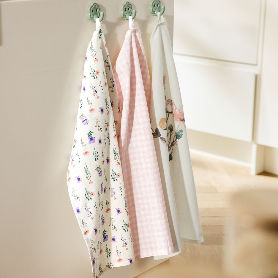 Trys raštuoti virtuviniai rankšluosčiai kabo ant kabliukų prie šviesios sienos.