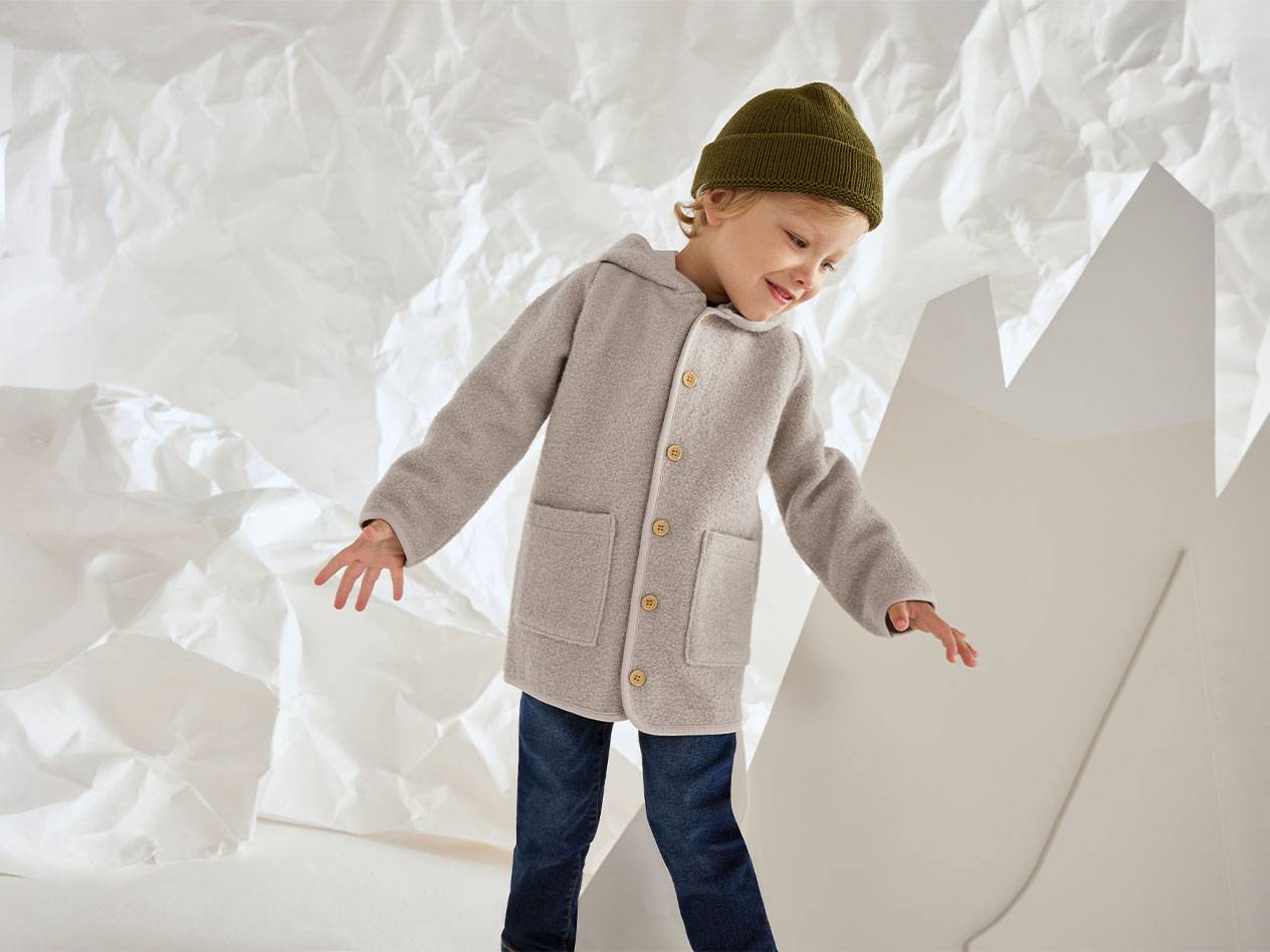 Berniukas su pilku vilnoniu švarku ir žalia kepure, besišypsantis raukšlėto popieriaus fone.