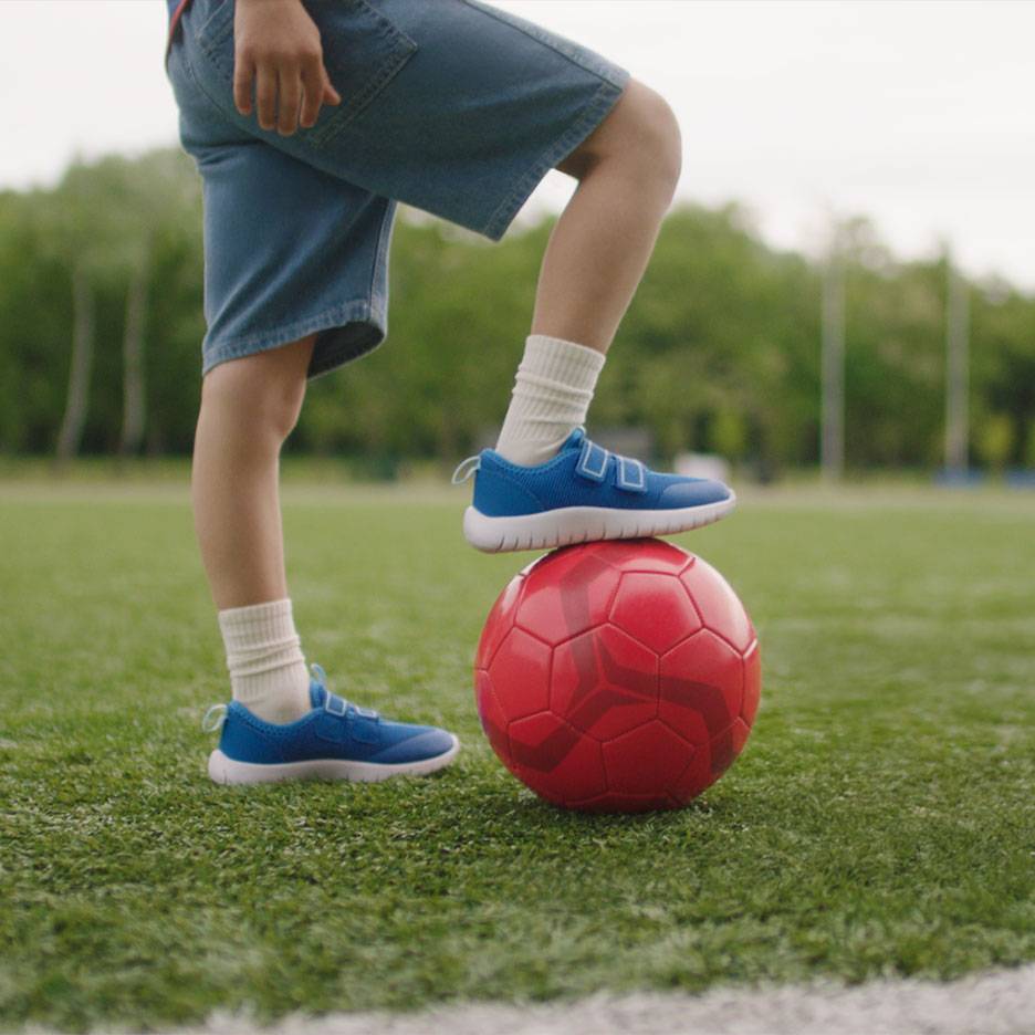 Vaikas su šortais ir mėlynais sportbačiais, viena koja ant raudono futbolo kamuolio.