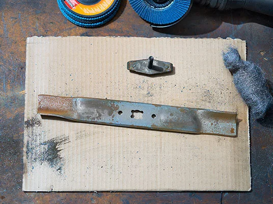 Rūdytas vejapjovės peilis ant kartono su abrazyviniais diskais ir plienine vata.