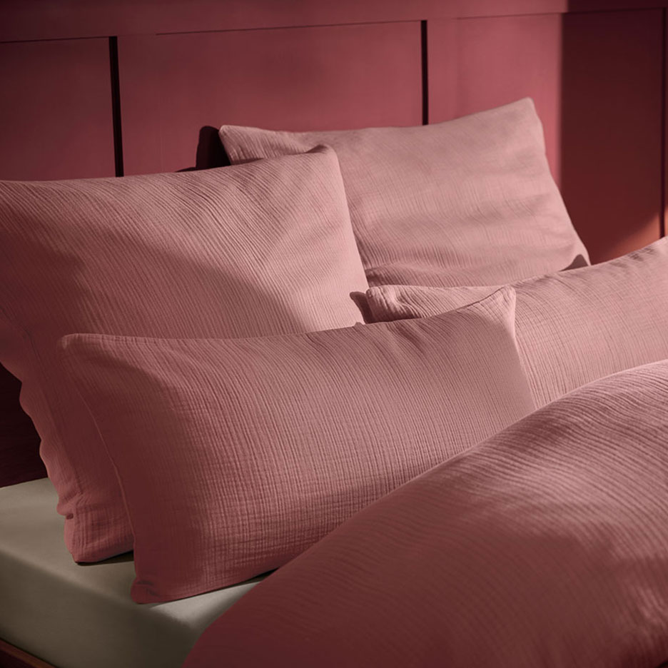 Rožinė pagalvė ir muslino patalynė ant lovos su mediniu galvūgaliu.