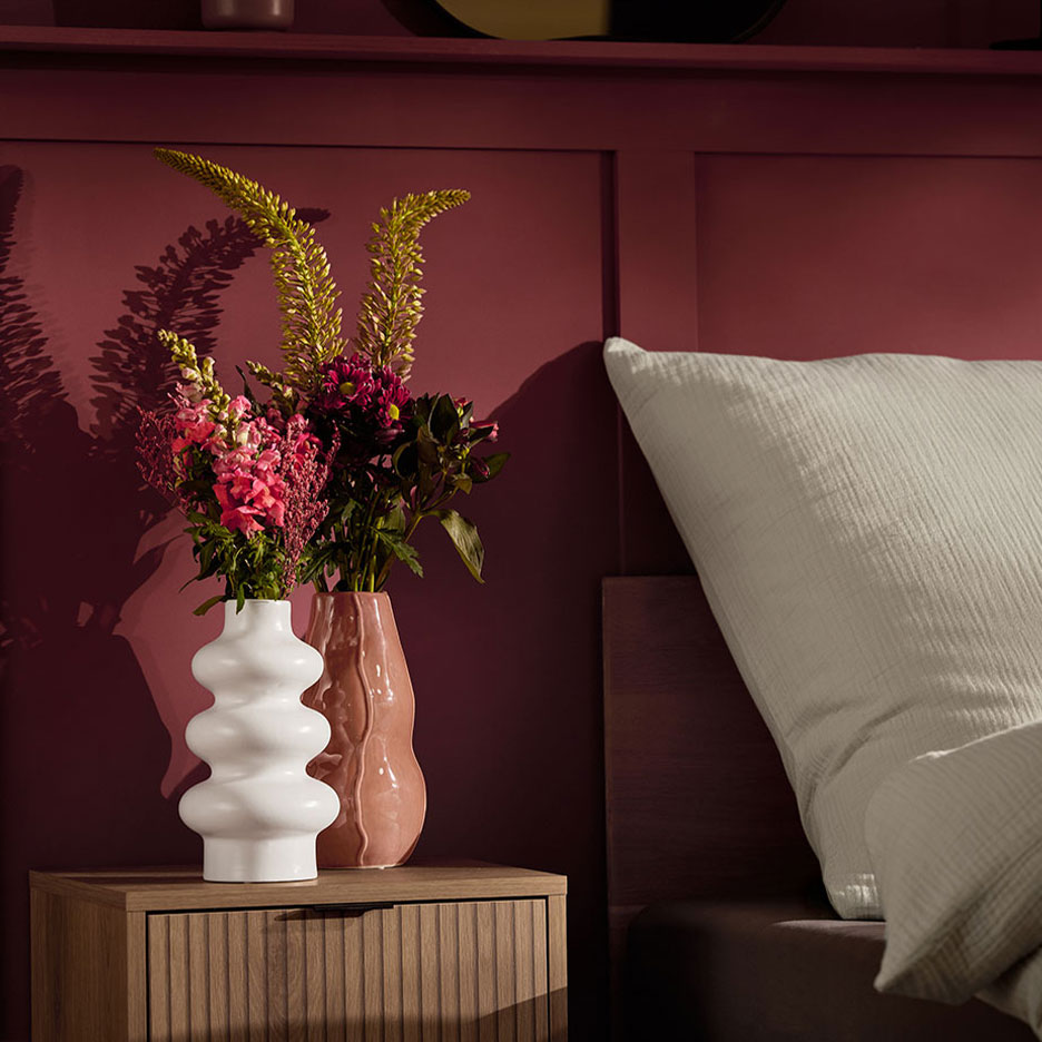 Dvi vazos su gėlėmis ant naktinio staliuko prie lovos su balta pagalve.