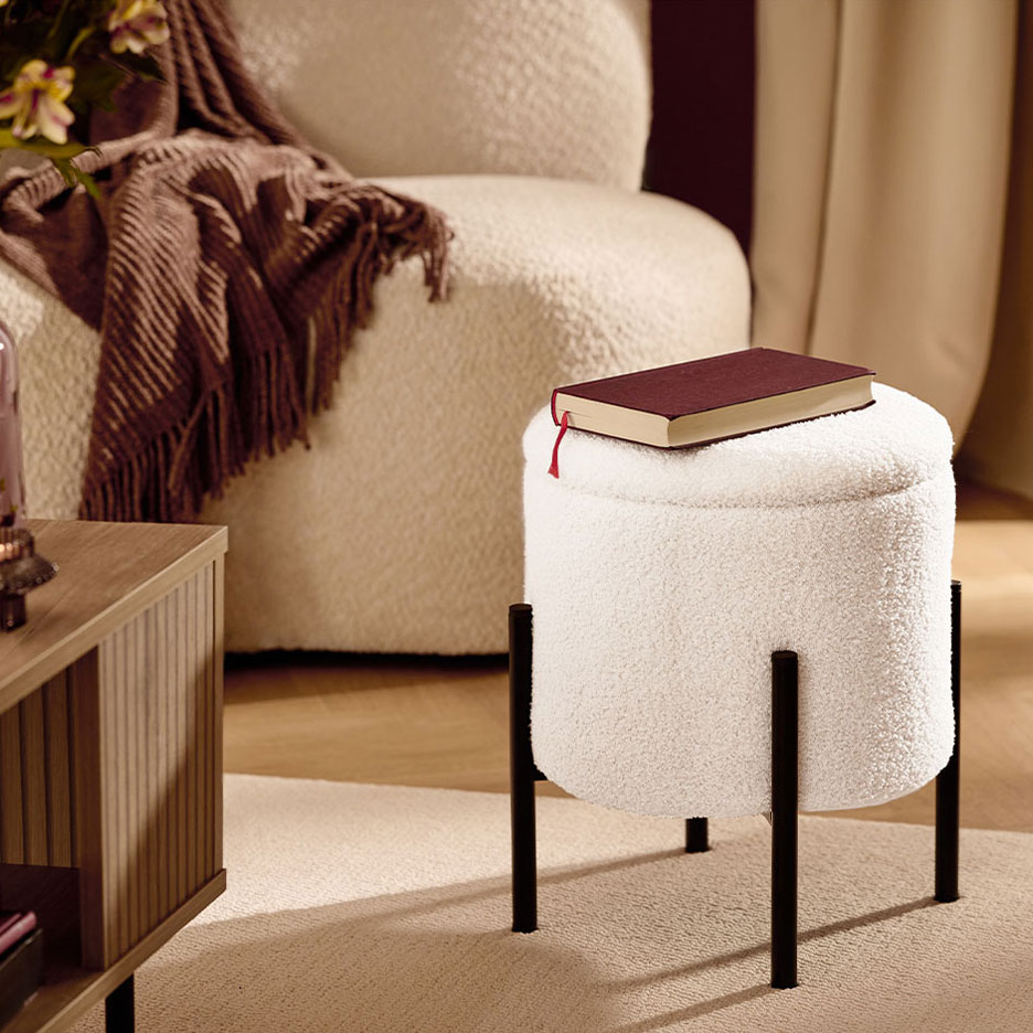 Baltas pūkuotas pufas su juodomis kojomis, raudona knyga, fotelis ir rudas pledas.