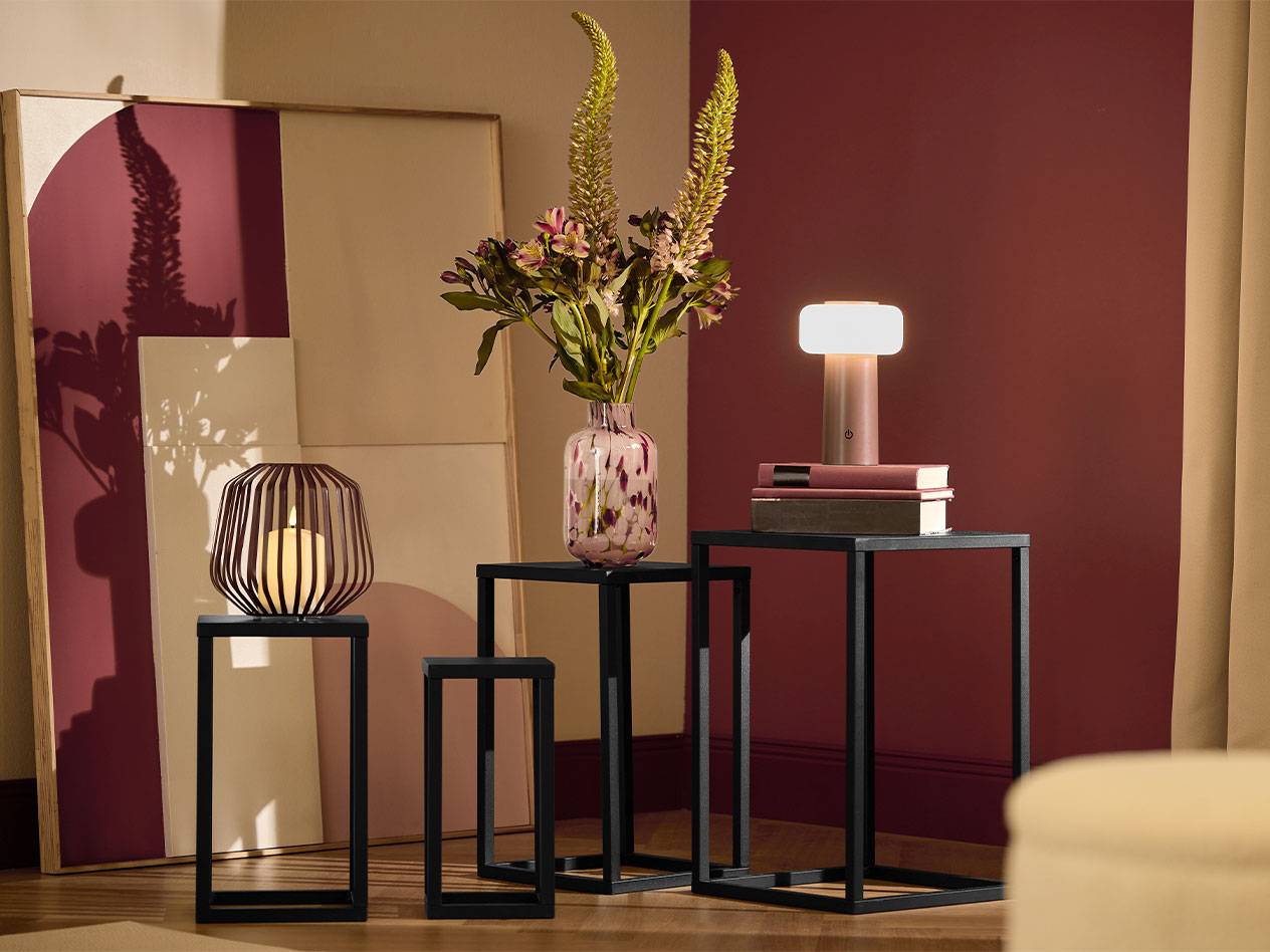 Trys juodi šoniniai staliukai su žvake, gėlių vaza ir lempa.