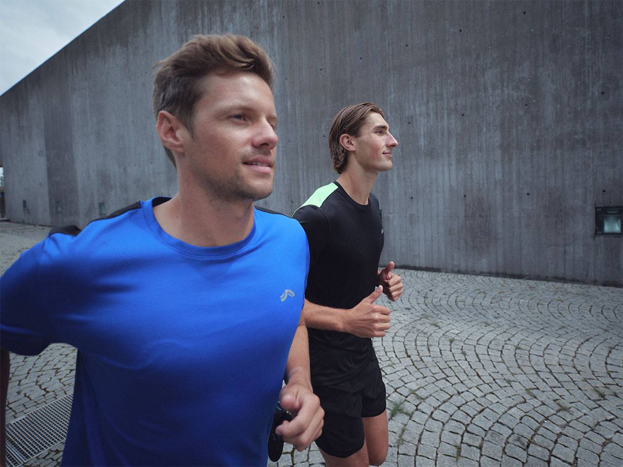 Du vyrai bėga su sportiniais marškinėliais ir šortais, vienas mėlynas, kitas juodas.