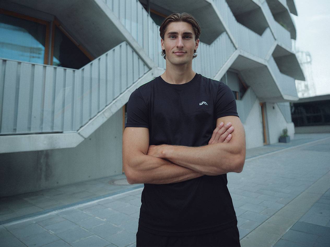 Vyras su juodais marškinėliais su logotipu ir juodais šortais, stovintis lauke sukryžiavęs rankas.