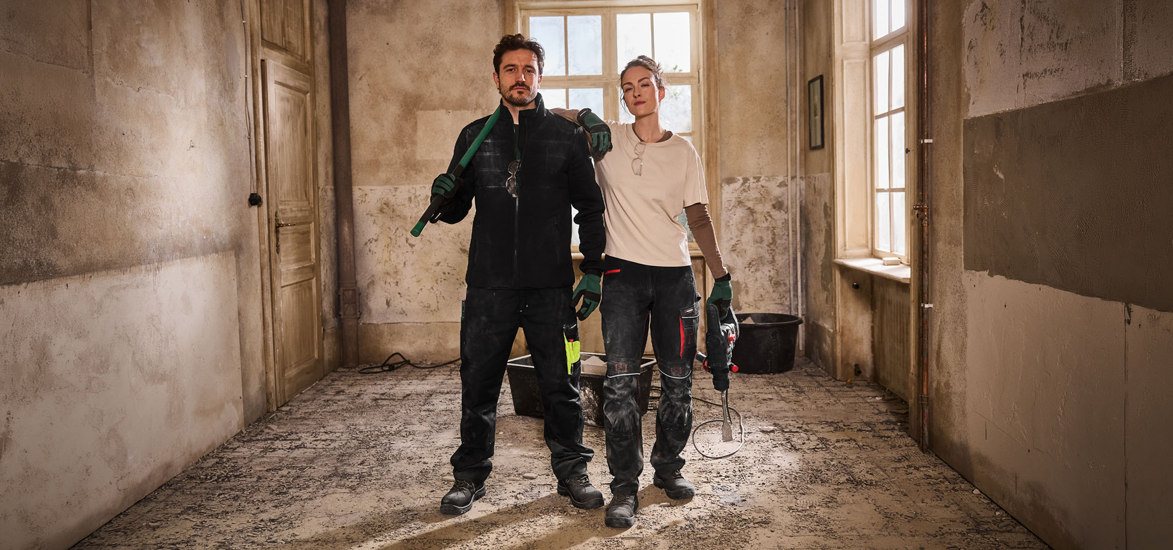 Du statybos darbuotojai su darbo drabužiais ir įrankiais statybvietėje.