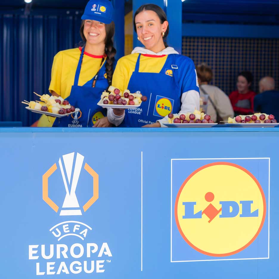 Dvi besišypsančios moterys su Lidl prijuostėmis siūlo vaisius ant iešmelių UEFA Europos lygos renginyje.