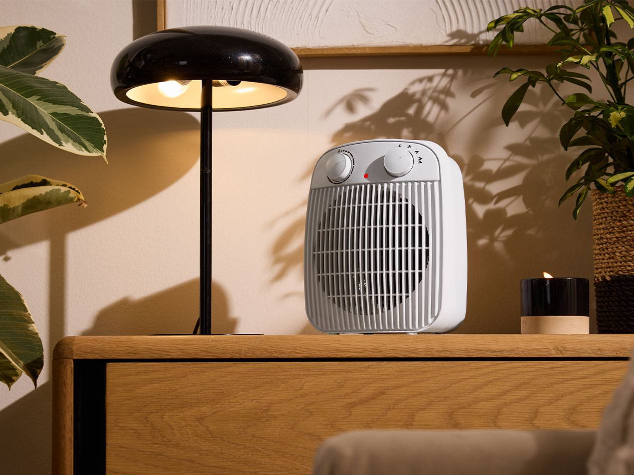 Baltas šildytuvas ir juodas stalo šviestuvas ant medinių baldų jaukioje patalpoje.