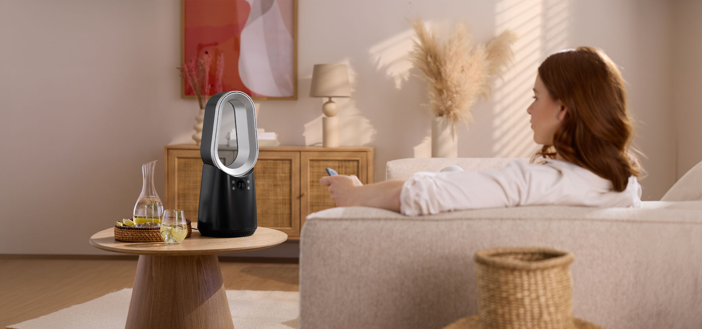 Moteris ant sofos su nuotolinio valdymo pulteliu, šalia modernus ventiliatorius ir gėrimai.