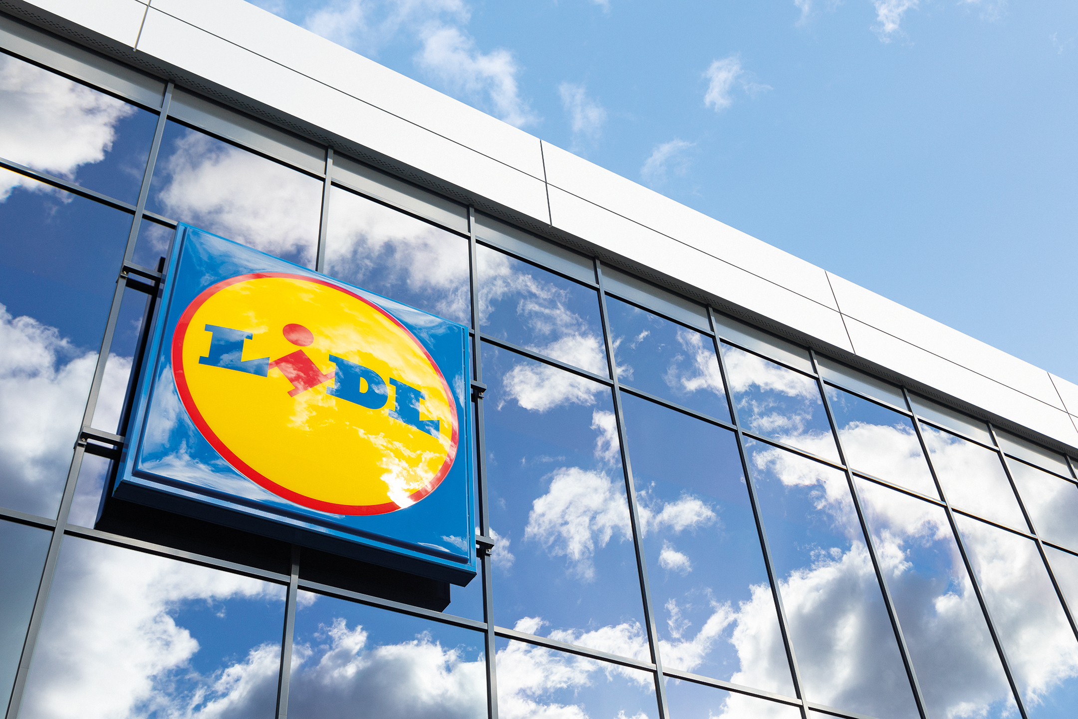 Lidl ženklas ant stiklinio pastato fasado, atsispindint dangui ir debesims.
