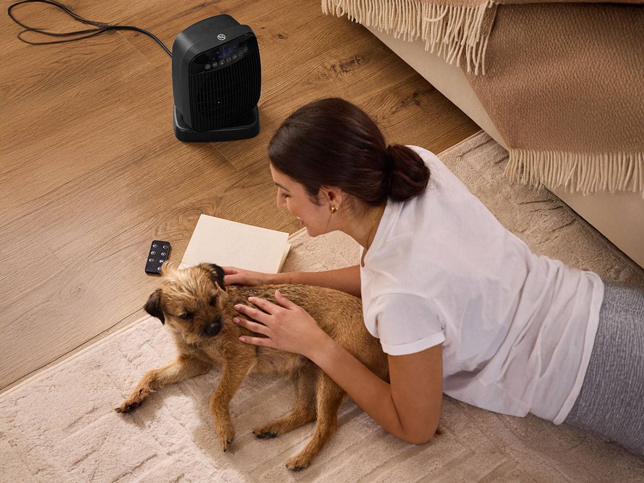 Moteris glosto šunį šalia juodo šildytuvo ant grindų.