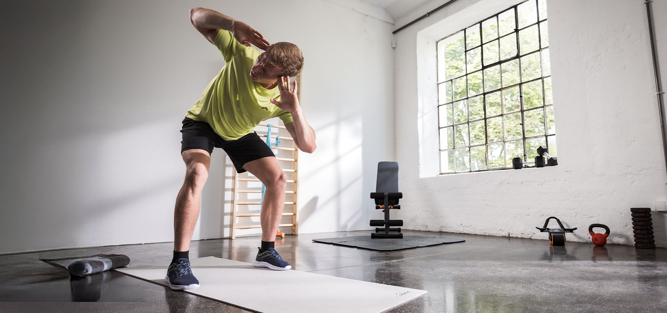Vyras sportuoja sporto salėje, naudodamas jogos kilimėlį ir hantelius.