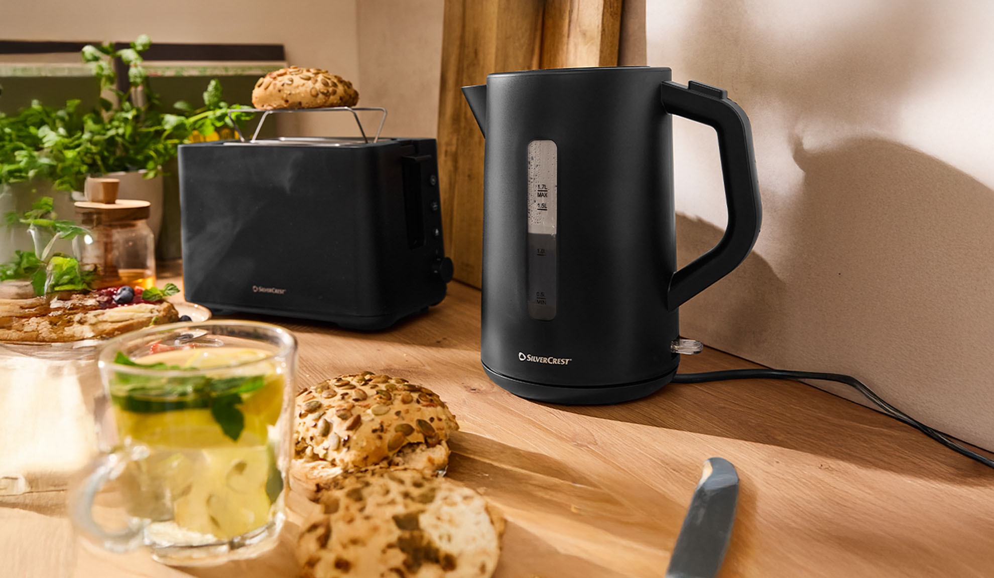 Juodas SilverCrest virdulys ir skrudintuvas ant medinio stalo su pusryčiais.