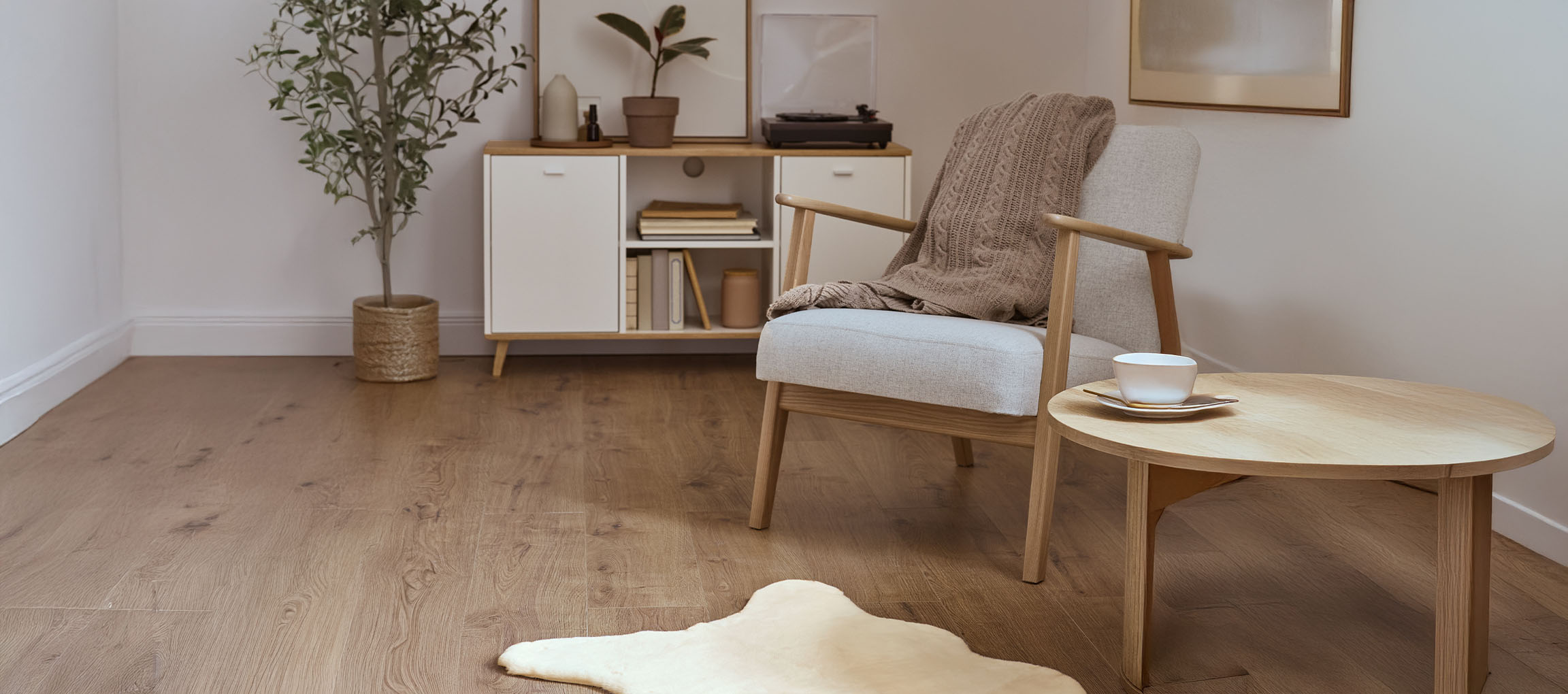 Pilkas audinio fotelis su megztu pledu, medinis kavos staliukas ir modernus bufetas.