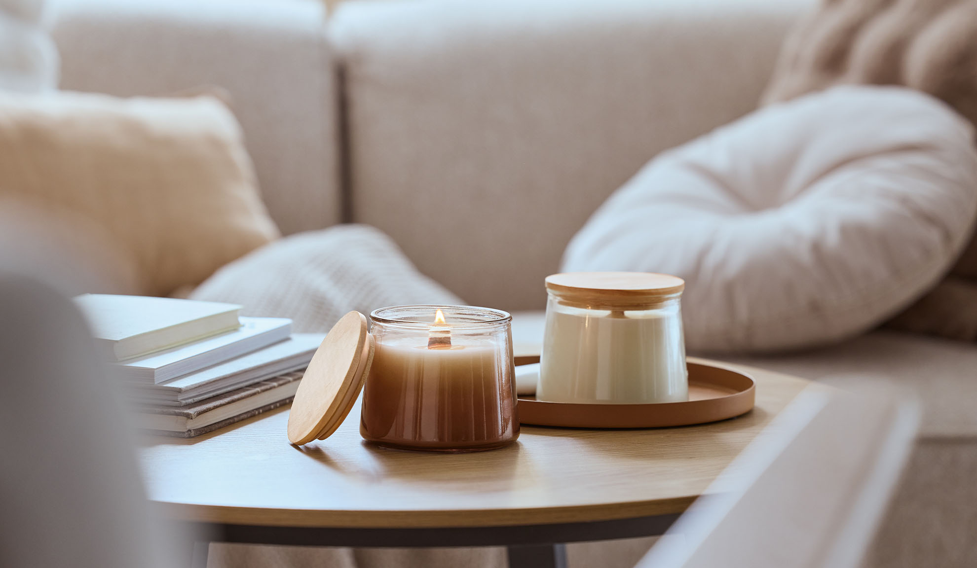 Dvi kvapiosios žvakės ant medinio stalo, viena dega, fone jaukus sofa.