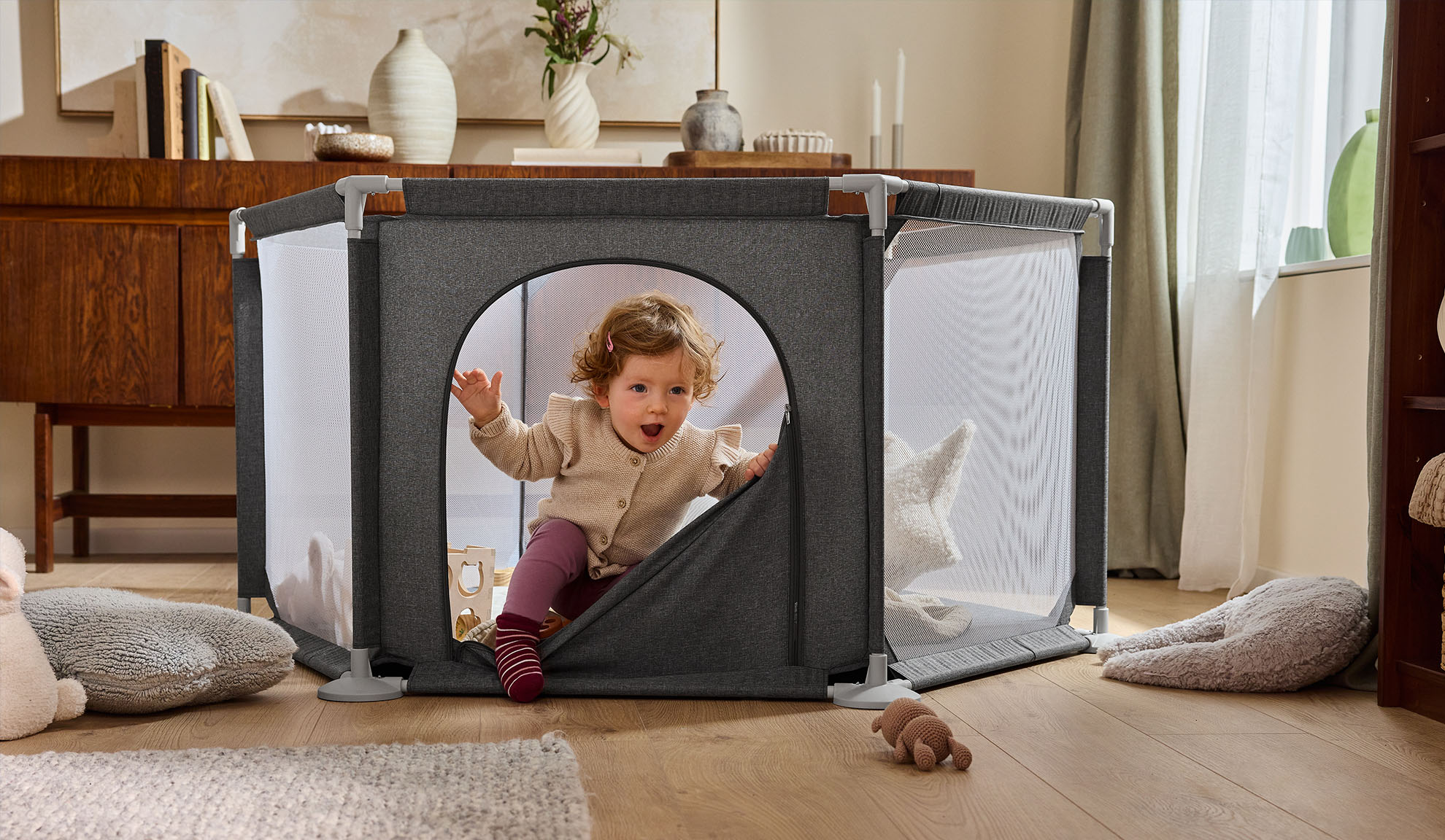 Laimingas kūdikis išropoja iš pilko maniežo jaukiame kambaryje.