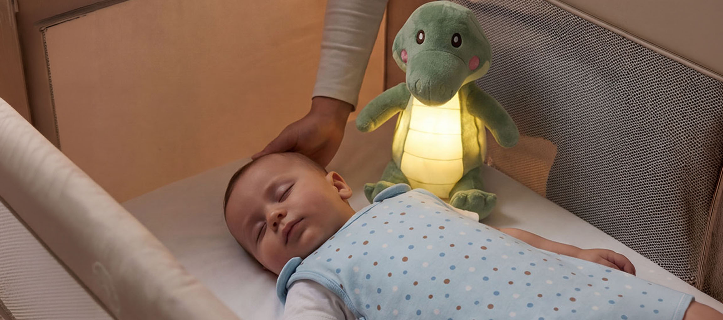 Kūdikis miega lovytėje su pliušiniu naktiniu šviestuvu, suaugęs glosto kūdikio galvą.
