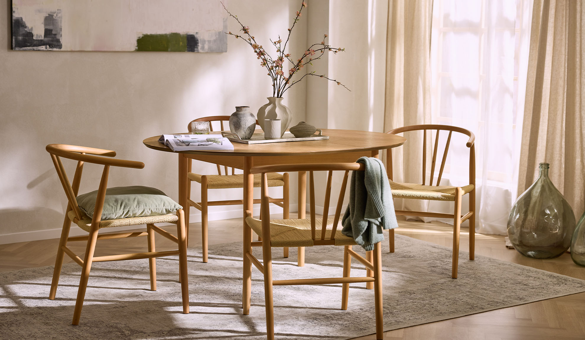Šviesi valgomojo zona su apvaliu mediniu stalu, medinėmis kėdėmis su virvinėmis sėdynėmis ir gėlių dekoru.