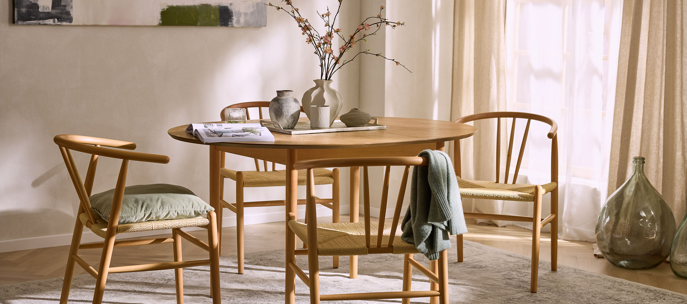 Valgomasis su apvaliu mediniu stalu, medinėmis kėdėmis su virvinėmis sėdynėmis ir dekoratyvinėmis vazomis.