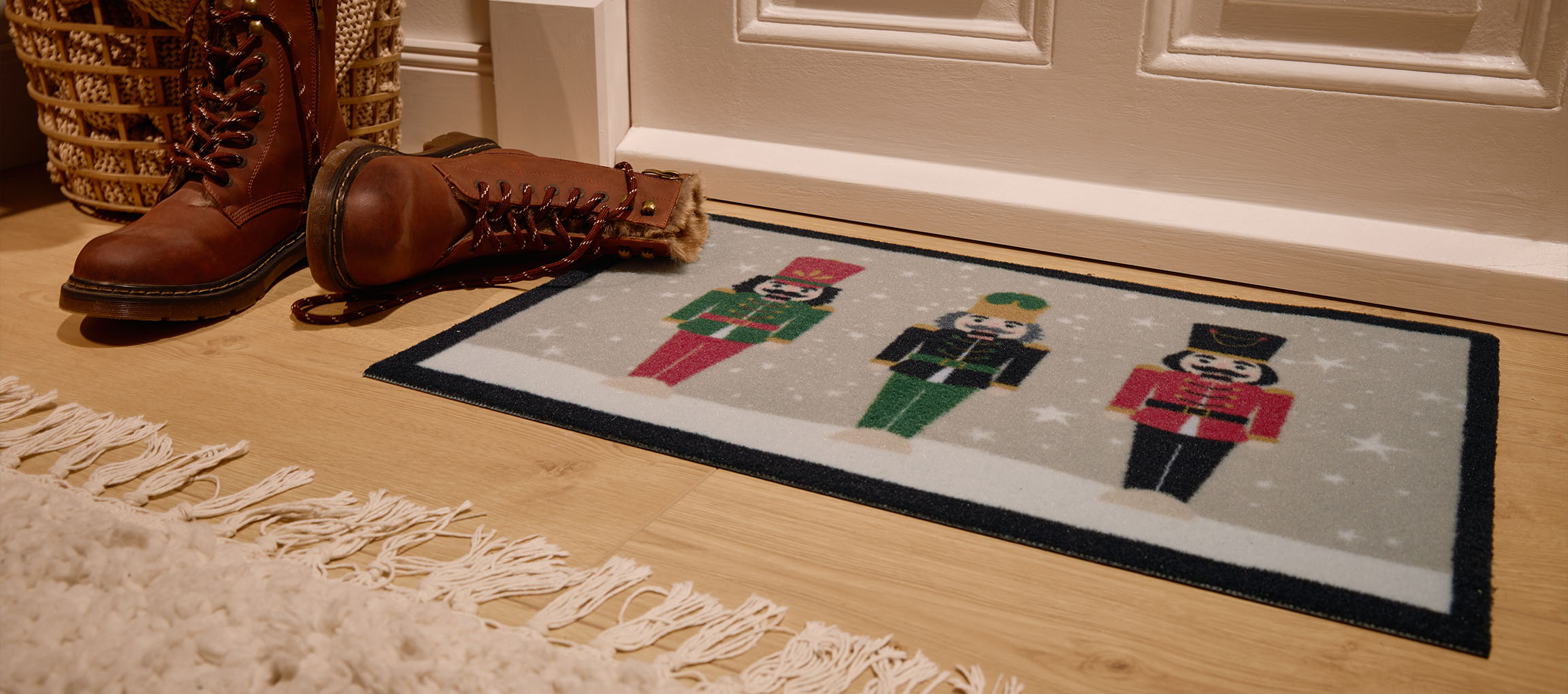 Kalėdinis durų kilimėlis su spragtukais, rudi batai ir pintas krepšys prie įėjimo.