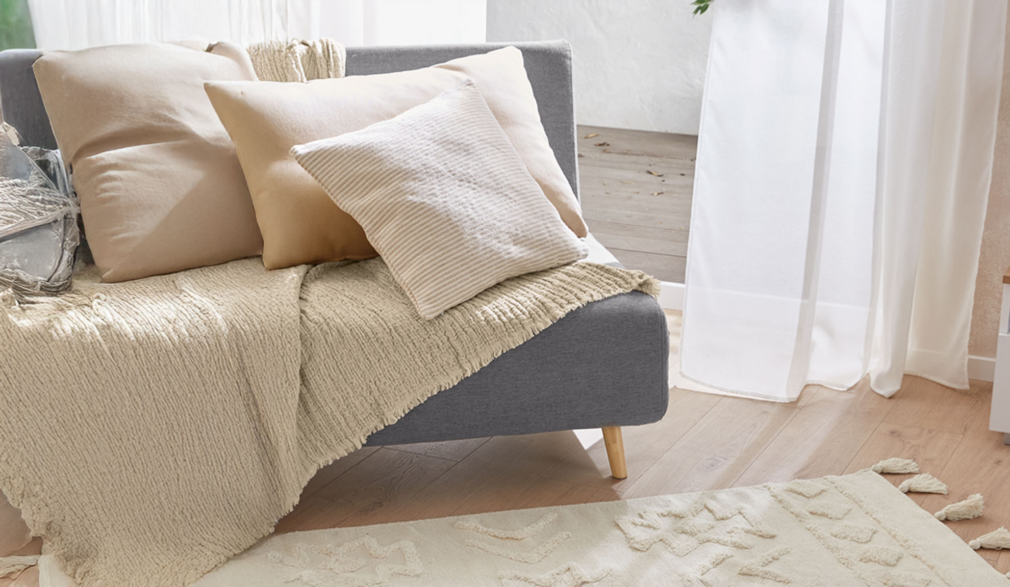 Pilka sofa su smėlio spalvos pagalvėmis ir tekstūruotu pledu šviesioje svetainėje.
