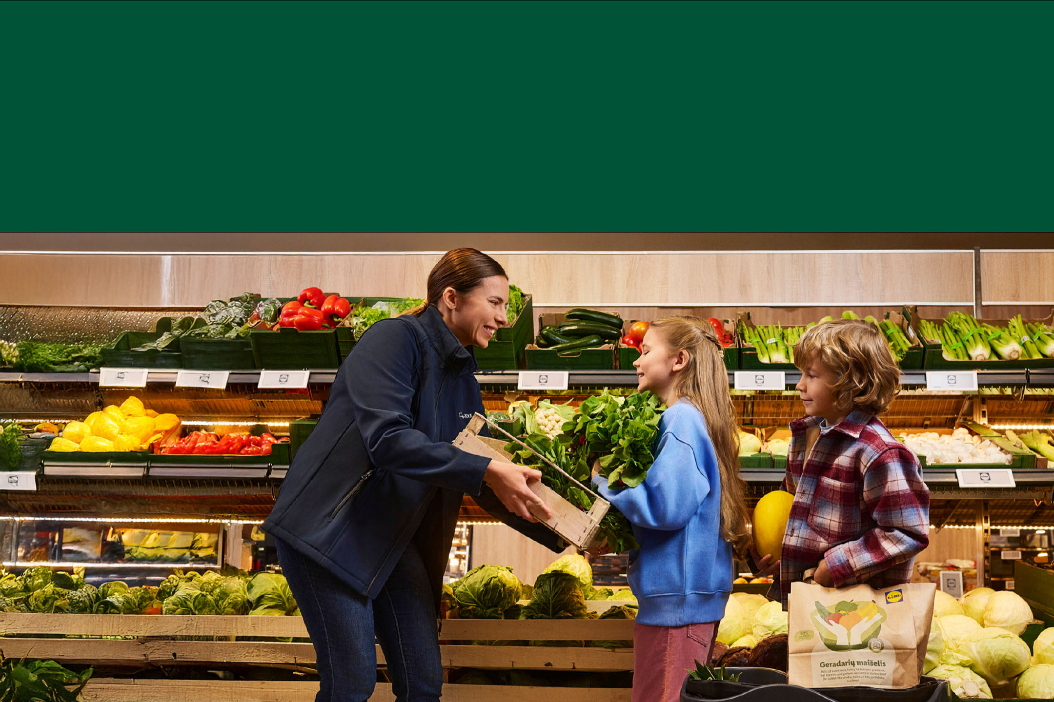 Moteris ir du vaikai parduotuvėje, apsupti šviežių daržovių, tokių kaip paprikos, cukinijos ir salotos.