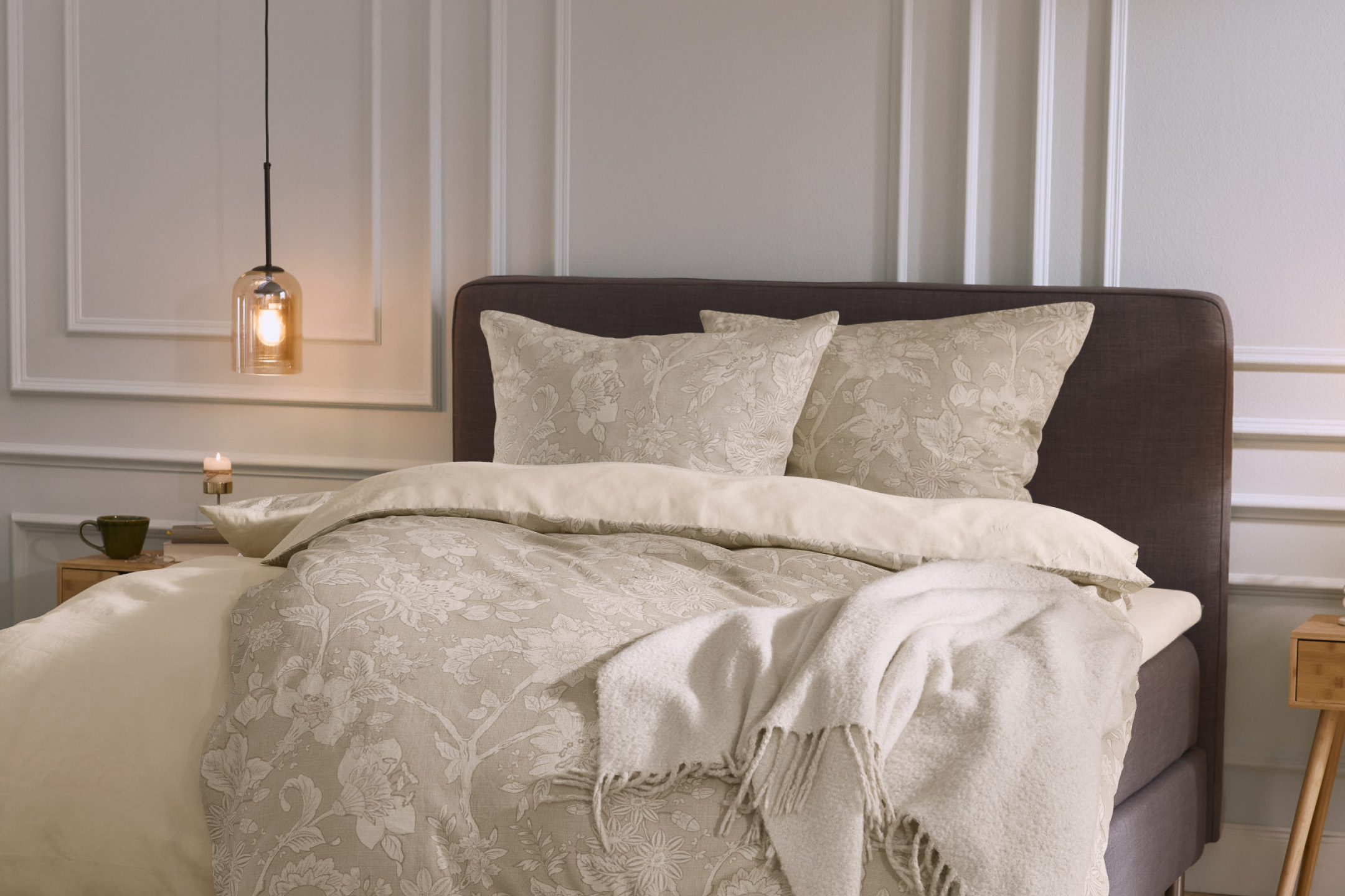 Šviesus miegamasis su gėlėta patalyne, tamsiai pilka lova ir šviesiu pledu.