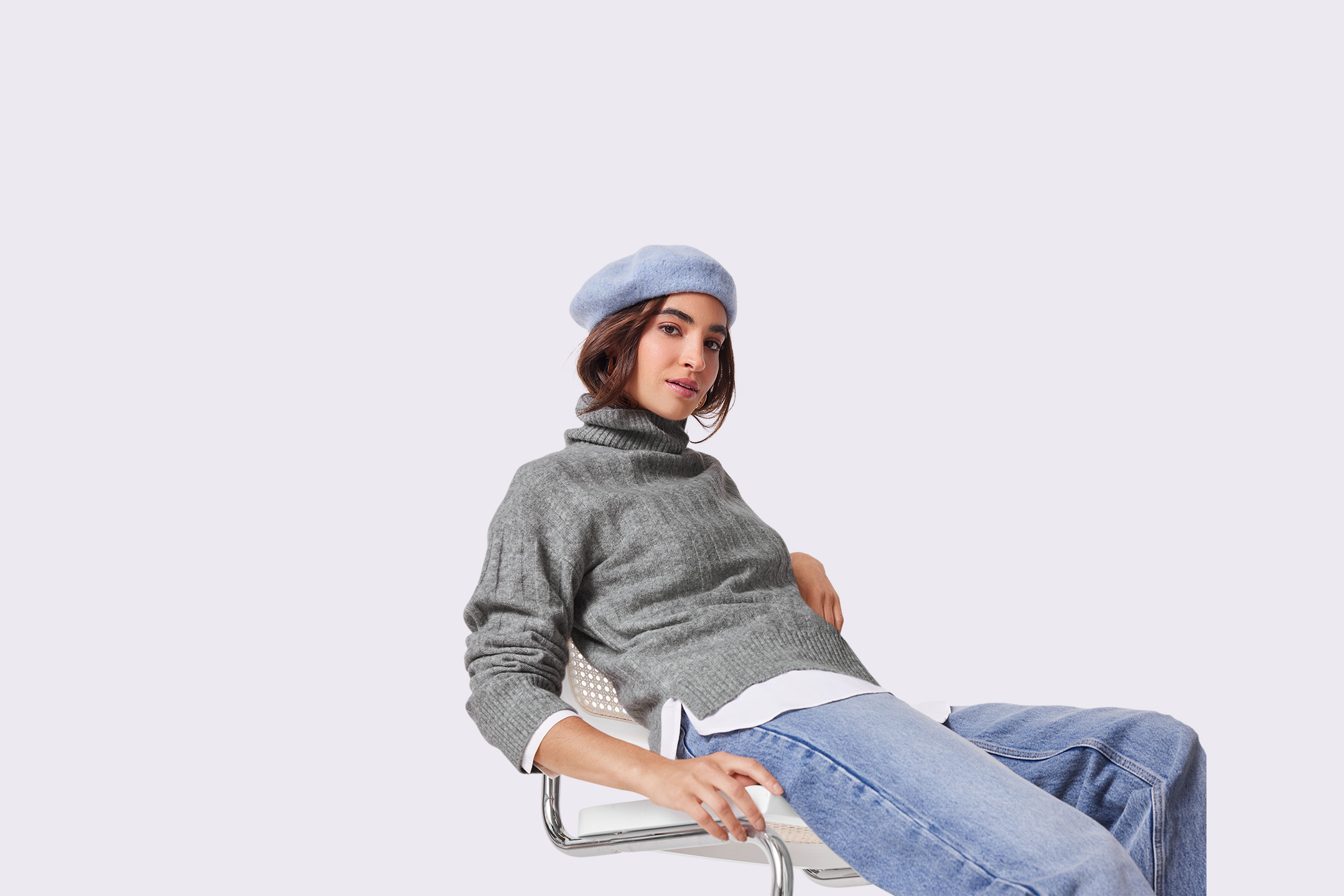 Moteris su pilku megztiniu, džinsais ir mėlyna berete sėdi ant kėdės.