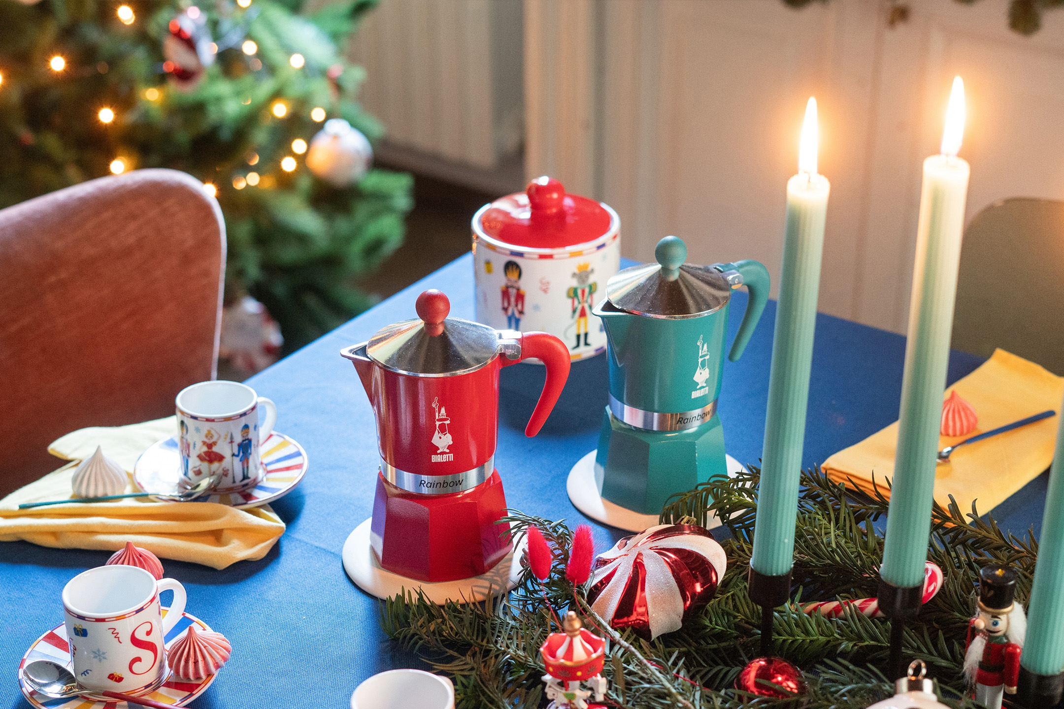 Raudoni ir turkio spalvos „Bialetti“ kavinukai, kalėdiniai puodeliai ir žvakės ant stalo.
