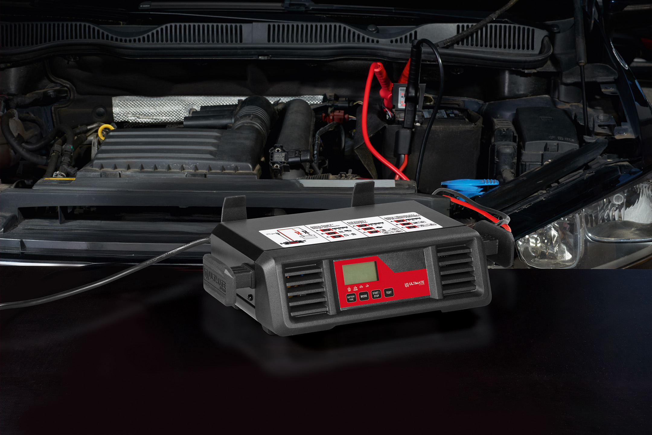 Automobilio akumuliatoriaus įkroviklis „Ultimate Speed“ su prijungtais gnybtais po atidarytu variklio gaubtu.