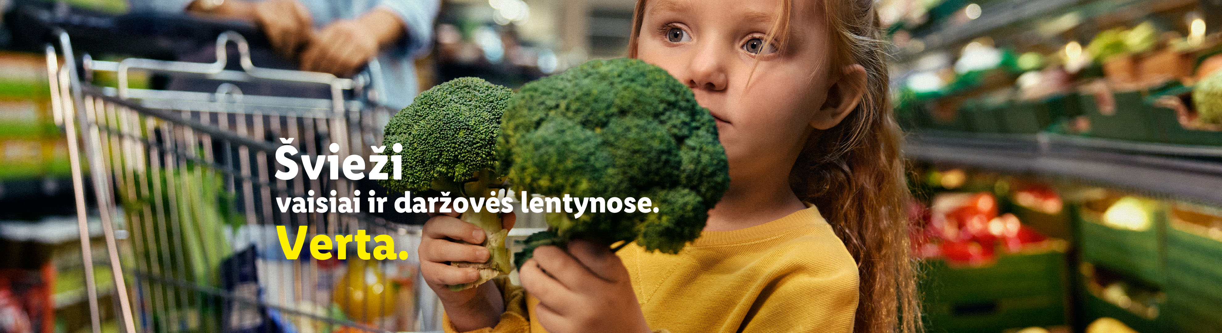 Mergaitė laiko brokolius, fone – prekybos vežimėlis ir švieži vaisiai bei daržovės.