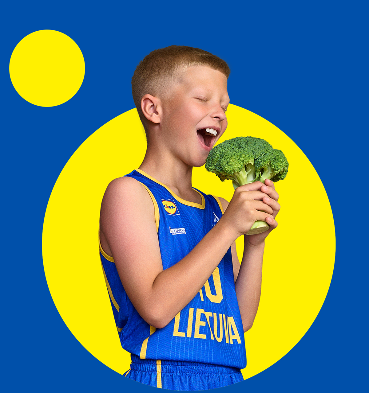 Berniukas mėgaujasi brokoliu, apsirengęs mėlynais sportiniais marškinėliais su geltonais akcentais.