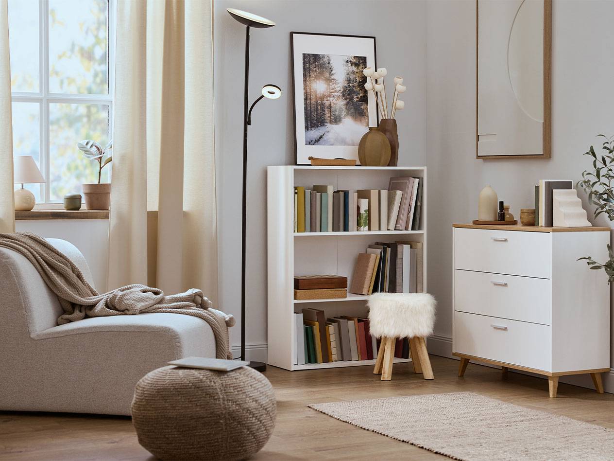 Jaukus svetainės kambarys su foteliu, knygų lentyna ir komoda neutraliomis spalvomis.