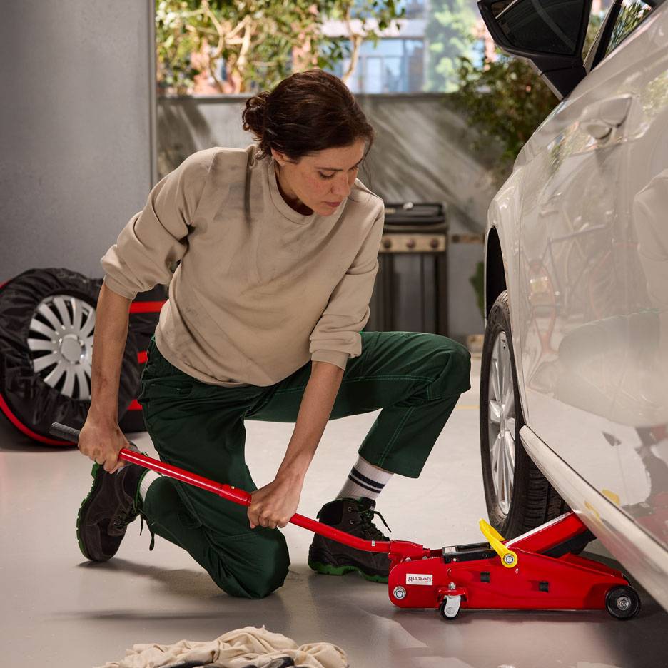 Moteris naudoja raudoną hidraulinį domkratą baltam automobiliui pakelti, šalia atsarginis ratas.