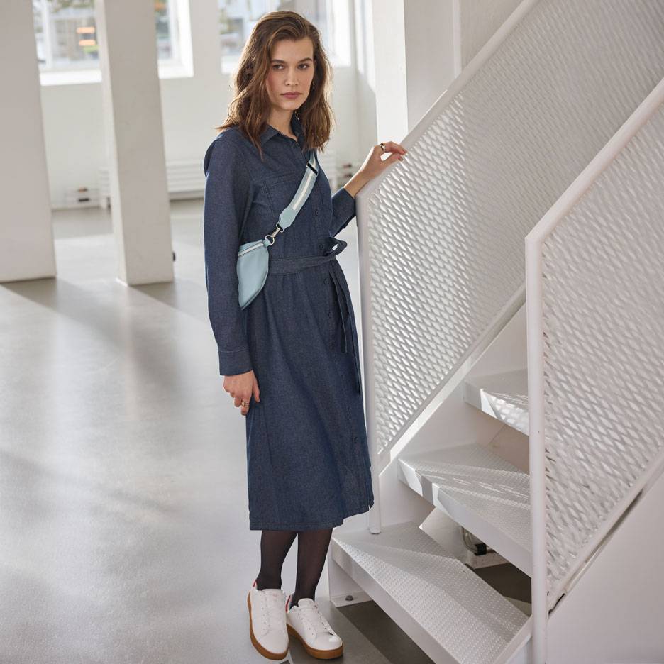 Moteris su džinsiniu marškinių tipo suknele, juodomis pėdkelnėmis ir baltais sportbačiais, su šviesiai mėlynu rankinuku.