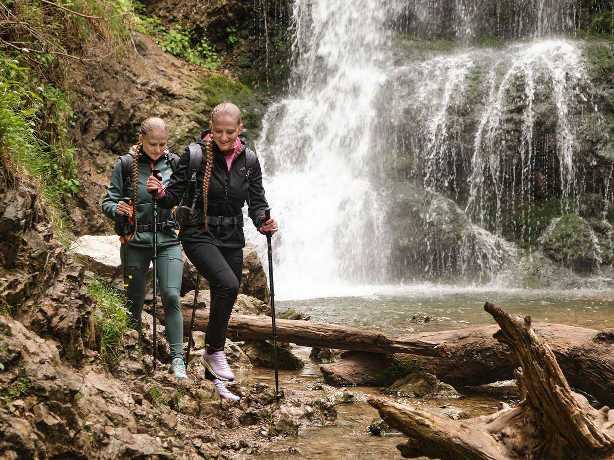 Dvi moterys su trekingo lazdom eina uolėtu taku prie krioklio.